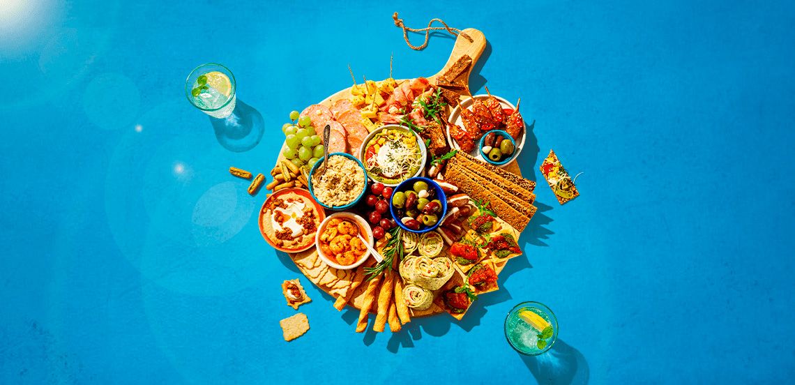 Borrelplank met diverse hapjes zoals kaas, vleeswaren, druiven, crackers, olijven, wraps en smeersels, geserveerd op een houten plank met twee glazen frisdrank op een blauwe achtergrond.