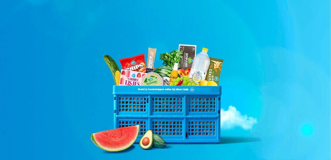 Blauwe Albert Heijn krat gevuld met diverse boodschappen zoals chips, water, komkommer, avocado, watermeloen, fruit, crackers, ijsjes, hummus en fles limonade tegen een blauwe achtergrond.
