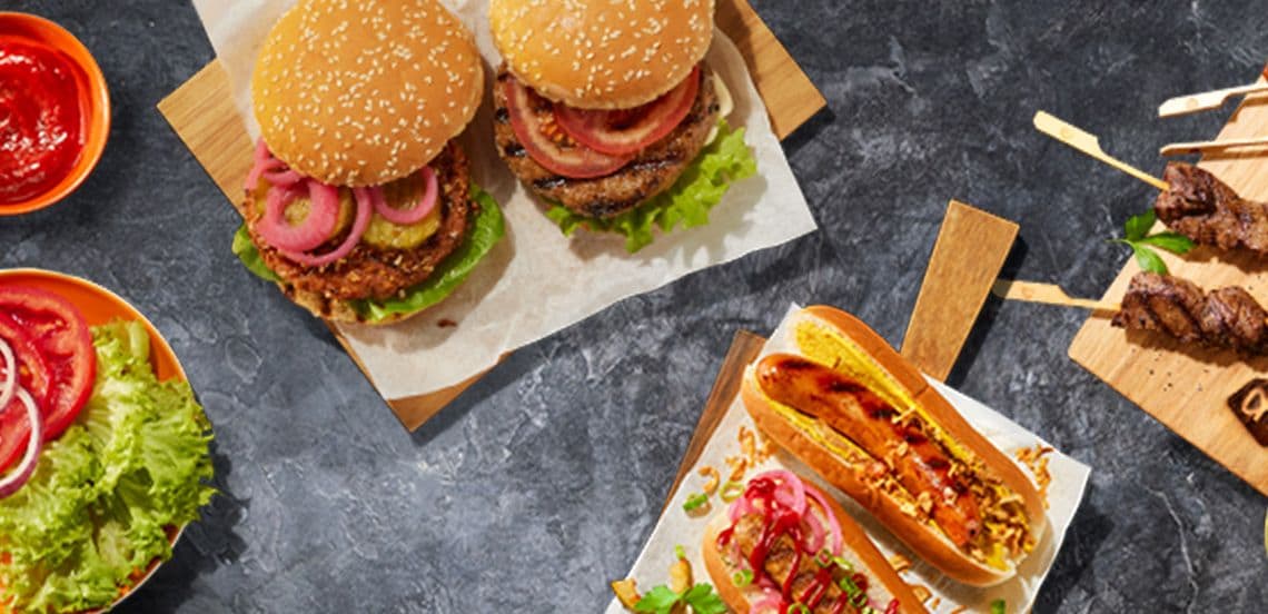 Twee hamburgers met sla, uien en augurk op een broodje, een hotdog met mosterd en ketchup, een broodje met groenten en twee spiesjes gegrild vlees op een donkere ondergrond, omringd door saus en verse groenten.