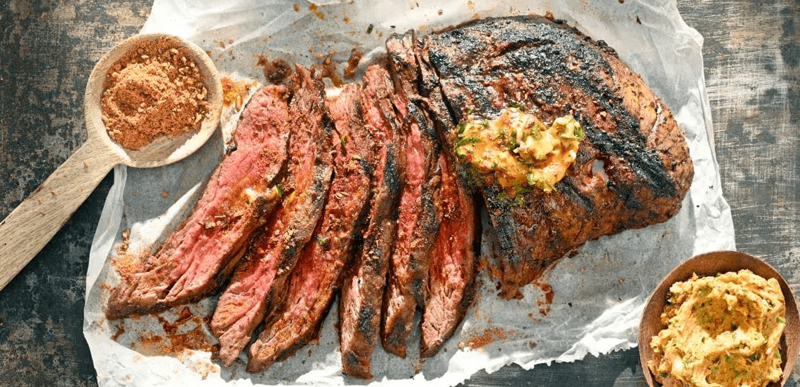 Gegrilde en in plakken gesneden steak met kruidenboter op bakpapier, met een houten lepel vol kruidenmix en een kom kruidenboter.