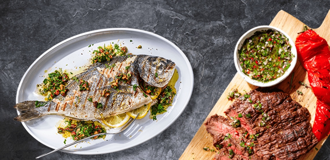 Gegrilde hele vis geserveerd met kruiden en citroen op een witte schaal, daarnaast gegrild rundvlees met groene chimichurri en geroosterde paprika op een houten plank.