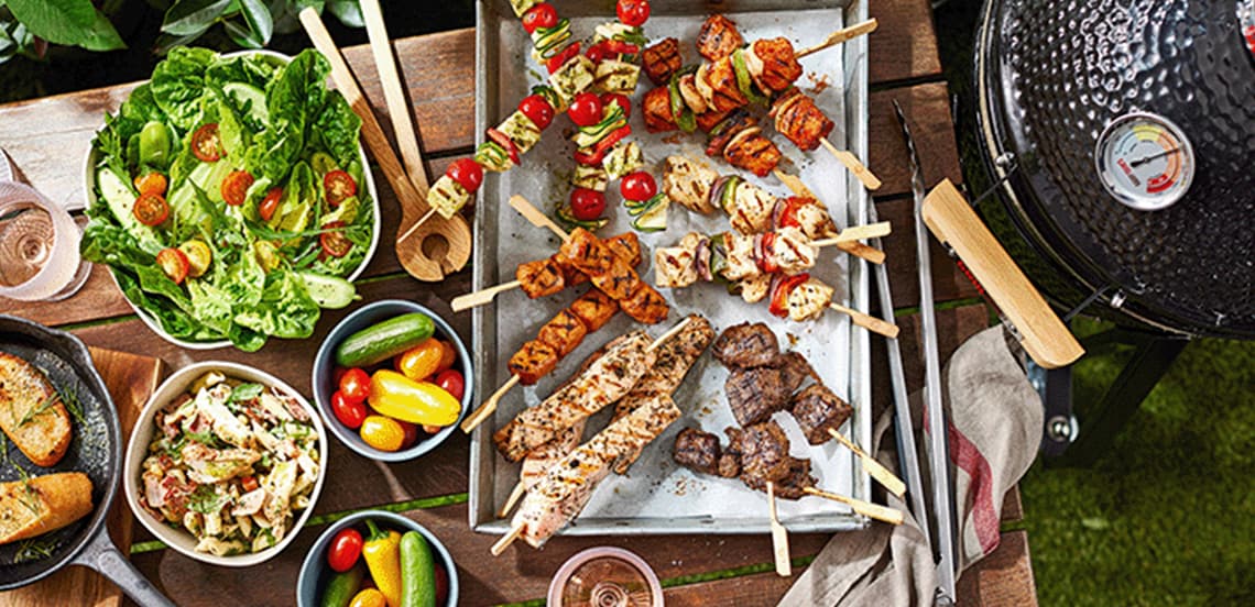 Barbecue met verschillende soorten gegrilde spiesen, verse groene salade, tomaatjes, brood en een pastasalade op een houten buitentafel naast een barbecue.