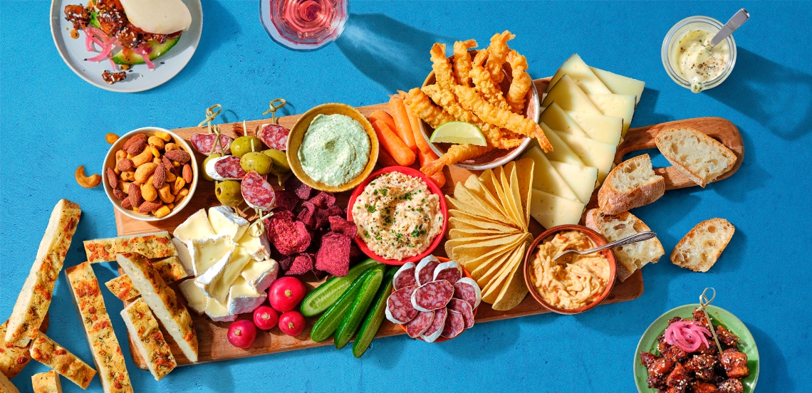 Borrelplank met diverse kazen, worst, groentesticks, toast, noten, smeersels, radijs, komkommer, groentechips, stokbrood en gefrituurde snacks op een blauwe ondergrond