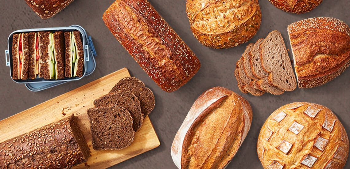 Verschillende soorten vers brood, waaronder volkoren, meergranen en bruin brood, deels gesneden en deels in hele broden, met een broodtrommel met sandwiches.