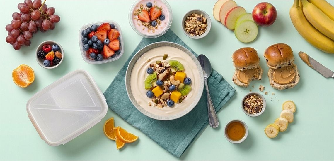 Verschillende soorten vers fruit zoals druiven, bessen, aardbeien, appels, sinaasappel en banaan, samen met een kom yoghurt met granola en fruit, ontbijtbroodjes met pindakaas, een bakje granola en honing, gepresenteerd op een lichtblauwe ondergrond.