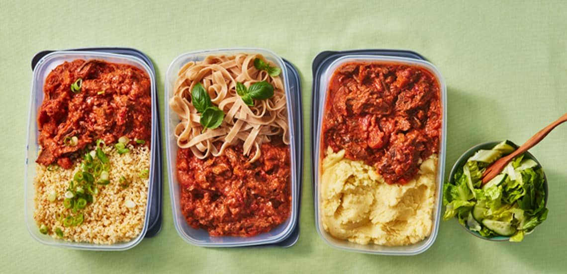 Drie maaltijdcontainers met stoofvlees in tomatensaus, geserveerd met respectievelijk couscous, tagliatelle en aardappelpuree, naast een kom gemengde salade met een houten lepel.