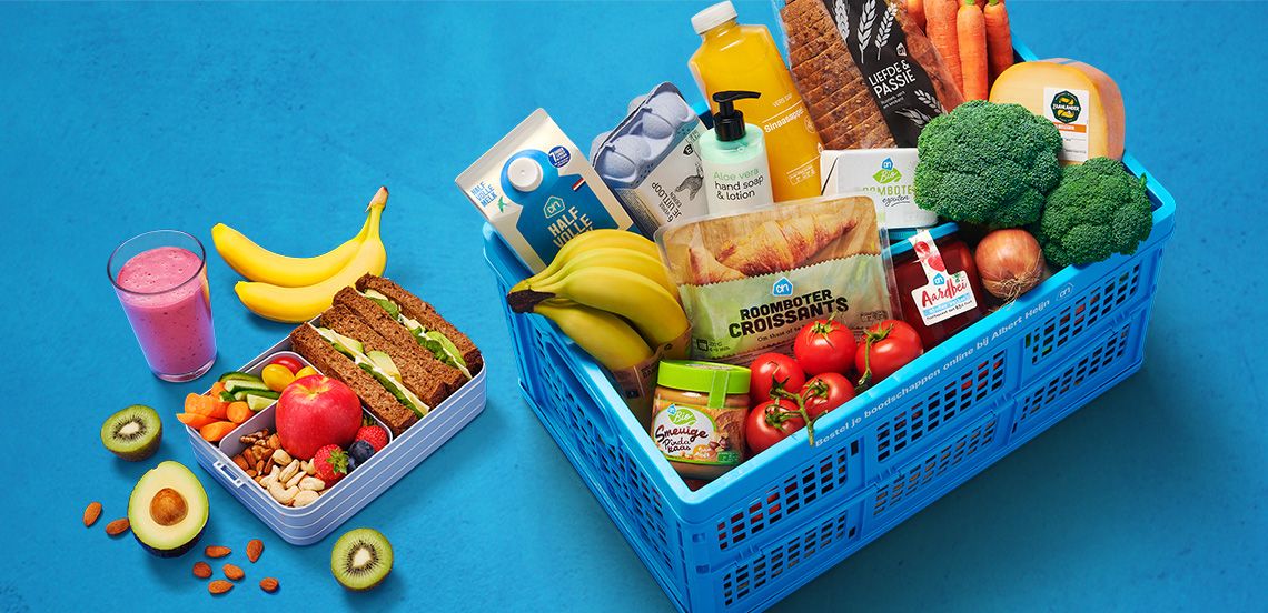 Albert Heijn blauwe winkelmand gevuld met boodschappen zoals brood, croissants, bananen, tomaten, broccoli, kaas, sinaasappelsap, halfvolle melk en handzeep, naast een lunchbox met volkoren sandwiches, fruit, noten en een smoothie op een blauwe ondergrond