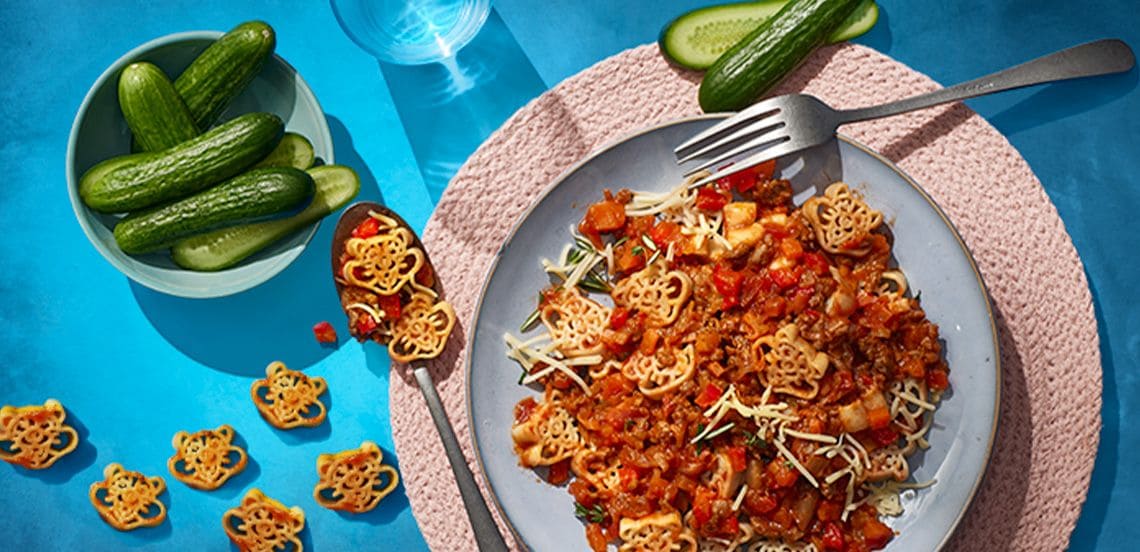 Kindervormpasta met pastasaus en geraspte kaas op een bord, geserveerd met komkommer op een blauw oppervlak