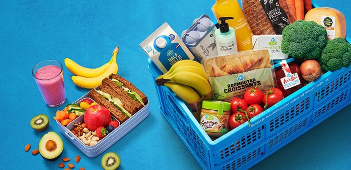 Blauwe boodschappenkrat van Albert Heijn gevuld met diverse verse producten zoals brood, bananen, melk, kaas, broccoli, tomaten, sinaasappelsap, croissants, eieren, handzeep en andere boodschappen; daarnaast een lunchbox met groente, fruit, noten, boterhammen en een glas roze smoothie.