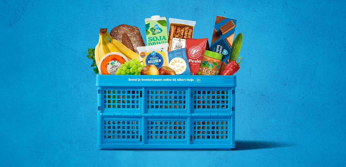 Blauwe Albert Heijn krat gevuld met diverse boodschappen, waaronder bananen, druiven, volkorenbrood, paprika, komkommer, kaas, melk, sojadrink, ontbijtgranen en koekjes, tegen een blauwe achtergrond.
