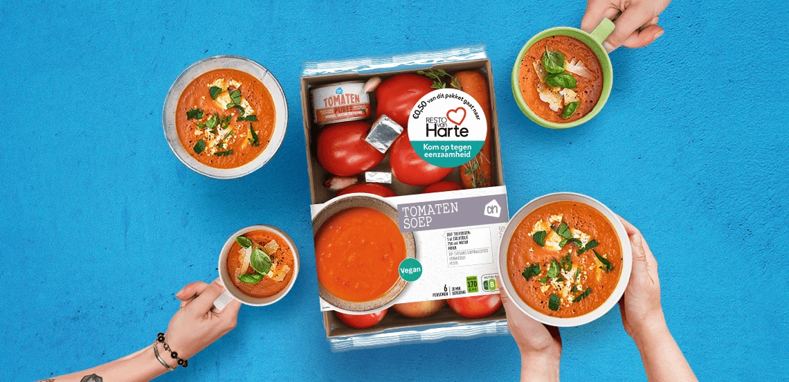 Voedselpakket van Albert Heijn voor tomatensoep met verse tomaten, tomatenpuree en bouillonblokjes, omringd door vier kommen tomatensoep die door verschillende handen worden vastgehouden tegen een blauwe achtergrond
