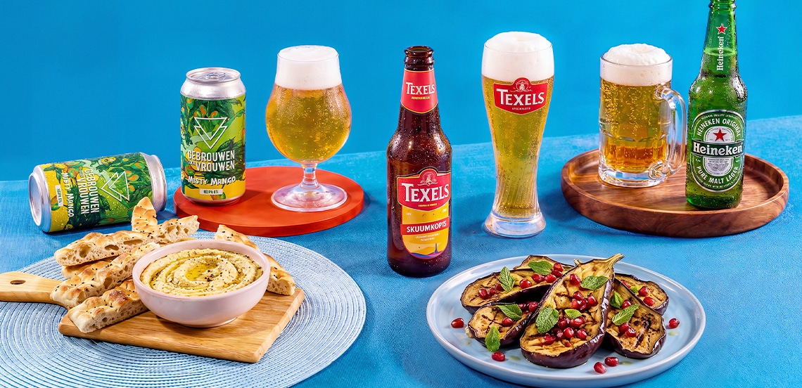 Diverse selectie van speciaalbieren, waaronder Gebrouwen door Vrouwen Misty Mango, Texels Skuumkoppe en Heineken, geserveerd met glaswerk, naast flatbread met hummus en gegrilde aubergine met granaatappelpitjes op een blauwe achtergrond.