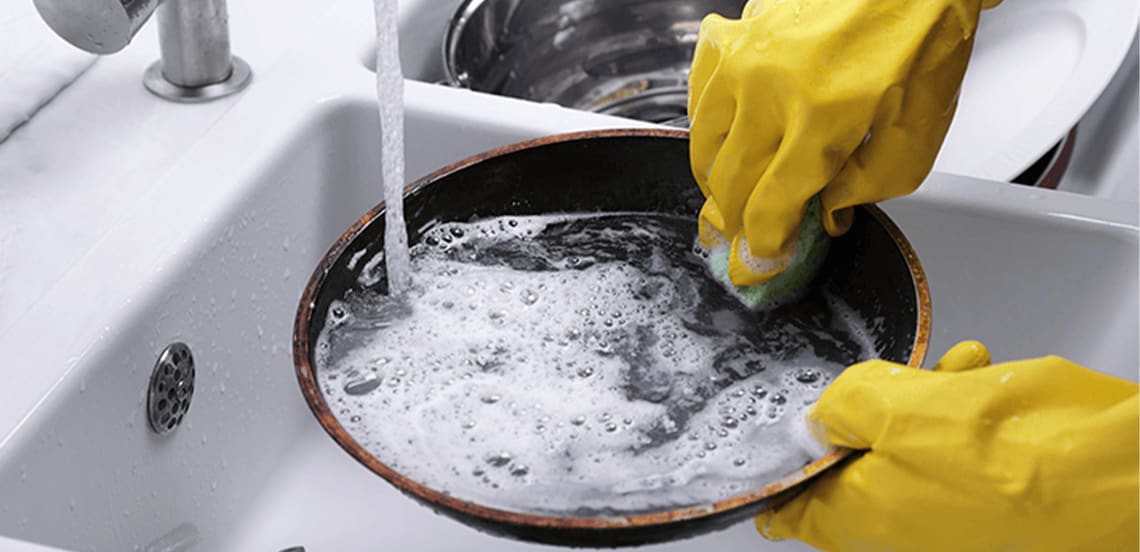 Iemand met gele schoonmaakhandschoenen wast een koekenpan met een spons onder stromend water in een gootsteen.