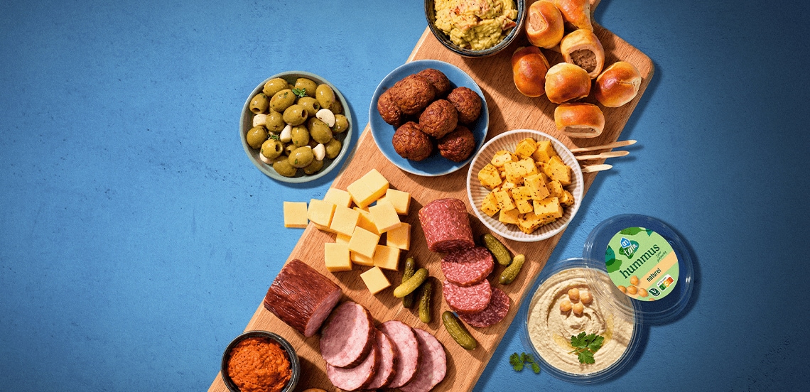 Borrelplank met diverse hapjes, waaronder groene olijven, kaasblokjes, salami, augurkjes, gehaktballetjes, kleine broodjes, guacamole, hummus van Albert Heijn en rode tapenade, geserveerd op een houten plank.
