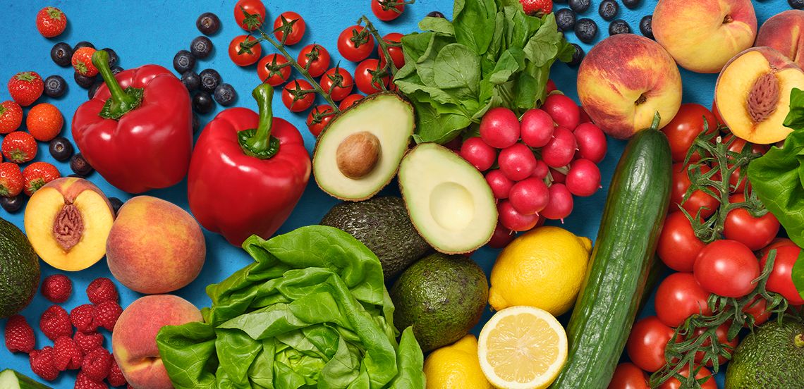 Verse groenten en fruit, waaronder rode paprika, avocado, tomaten, citroen, sla, perziken en bessen op een blauwe achtergrond.