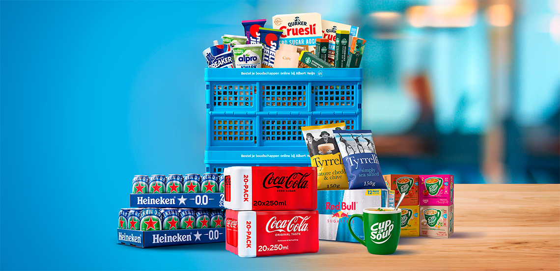 Blauwe boodschappenkrat gevuld met diverse A-merk producten zoals Quaker Cruesli, Breaker, Alpro, Heineken 0.0, Coca-Cola, Red Bull, Cup-a-Soup en Tyrrells chips, uitgestald op een houten tafel tegen een blauwe achtergrond.