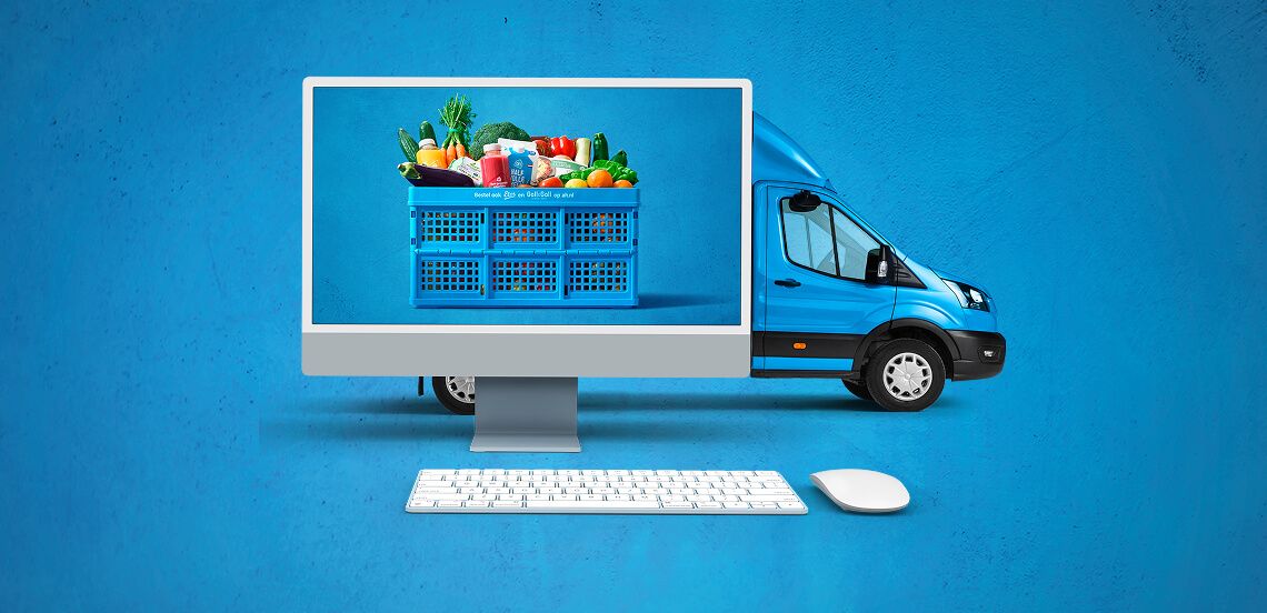 Computer met afbeelding van een blauwe Albert Heijn boodschappenkrat vol boodschappen, op de achtergrond een blauwe bezorgbus, toetsenbord en muis op een blauwe ondergrond