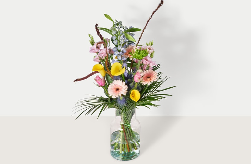 Boeket met diverse kleurrijke bloemen, waaronder gele calla lelies, roze gerbera’s en groene bladeren in een glazen vaas