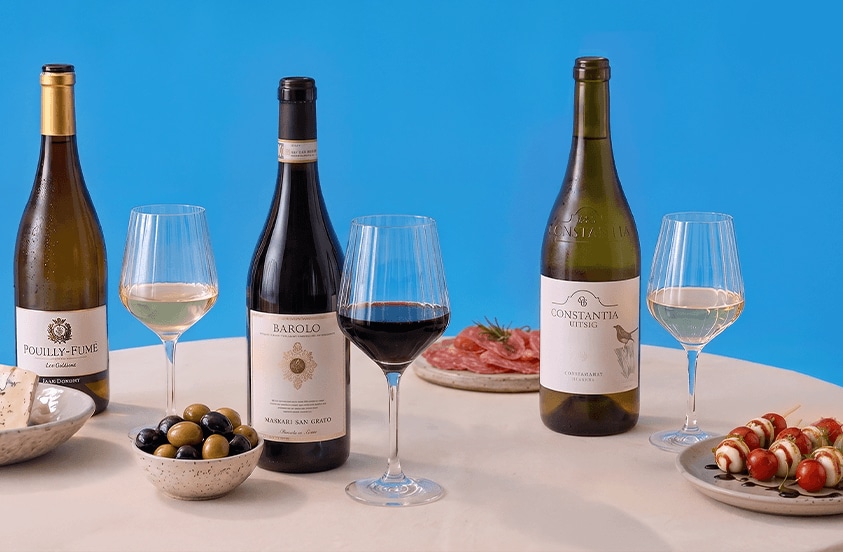Drie flessen wijn (Pouilly-Fumé, Barolo en Constantia Uitsig) met drie glazen wijn, een schaaltje olijven, een bord met charcuterie en een bord met spiesjes van mozzarella en tomaat, gepresenteerd op een tafel tegen een blauwe achtergrond.