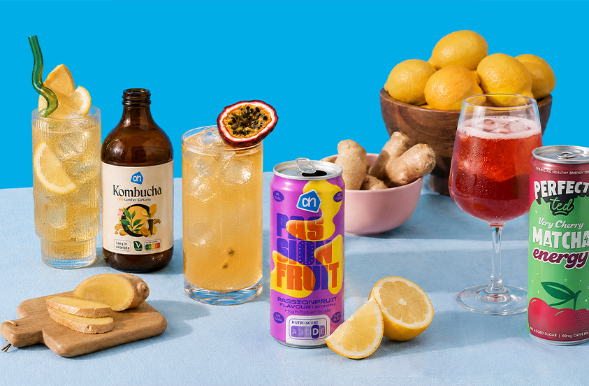 Een kleurrijke compositie van verfrissende dranken, waaronder AH Kombucha, een AH Passievrucht drank, Perfect Ted Very Cherry Matcha Energy, en diverse glazen met ijs, citroen, passievrucht en gember. Op de achtergrond staan een kom met citroenen en een bakje met gember, geschikt voor een zomerse sfeer.