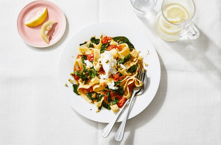 Tagliatelle met spinazie, tomaat en ricotta op een wit bord, geserveerd met citroenpartjes.