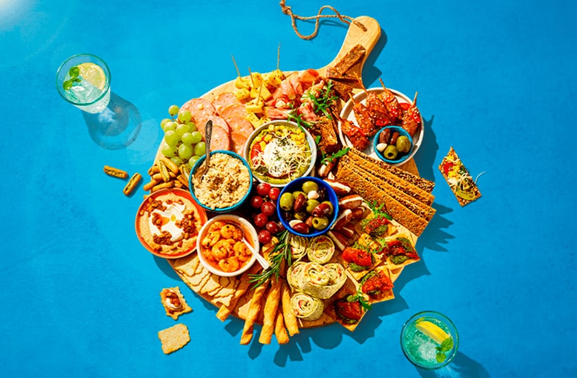 Borrelplank met diverse hapjes, waaronder kaasblokjes, vleeswaren, olijven, druiven, dips, wraps, toastjes en groenten, geserveerd op een houten plank boven een blauwe achtergrond.