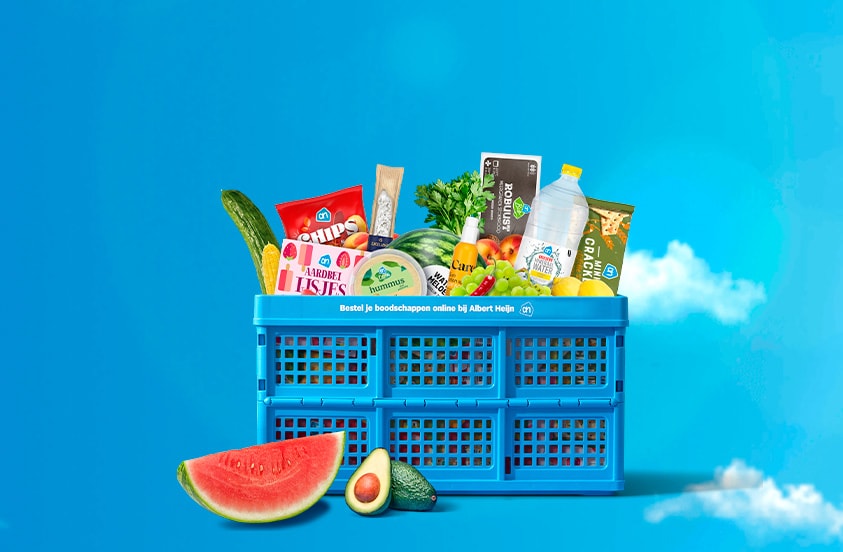 Blauwe Albert Heijn krat gevuld met diverse boodschappen, waaronder chips, aardbei ijssnoepjes, hummus, komkommer, watermeloen, avocado, crackers, een fles water, brood, druiven, appels en peterselie, geplaatst op een blauwe achtergrond met wolken.