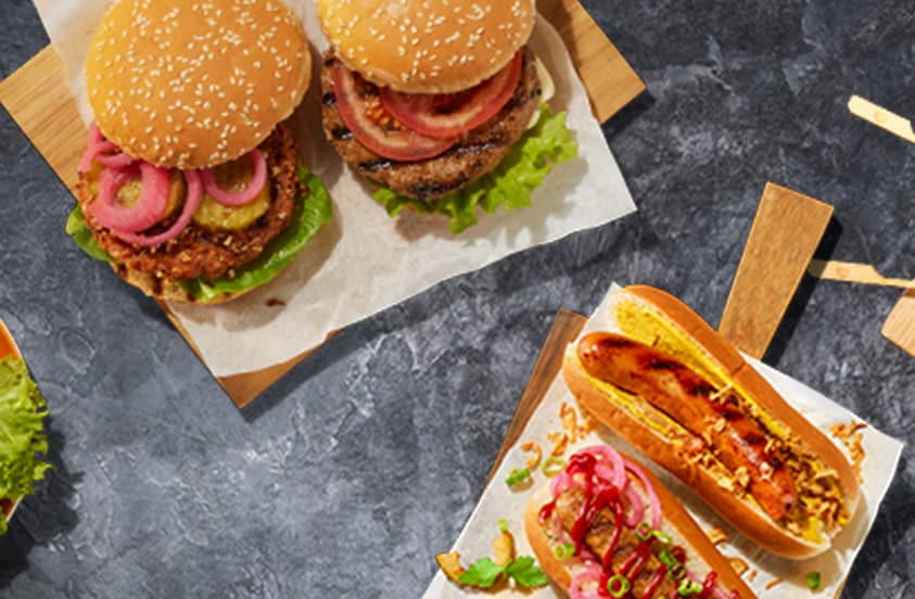 Twee hamburgers met sla, tomaat en ingelegde ui, en twee hotdogs met diverse toppings, geserveerd op houten planken.
