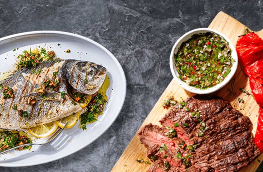 Gegrilde hele vis geserveerd met kruiden en citroen op een witte schaal, daarnaast gegrild rundvlees met groene chimichurri en geroosterde paprika op een houten plank.