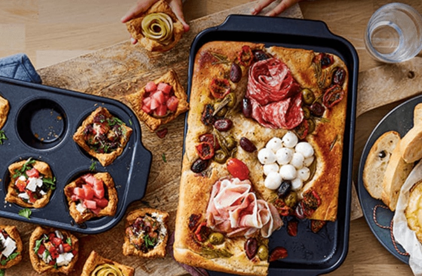 Gevulde borrelhapjes en focaccia met Italiaanse vleeswaren, mozzarella, olijven en groenten, geserveerd op een houten plank met een glas water en stokbrood.