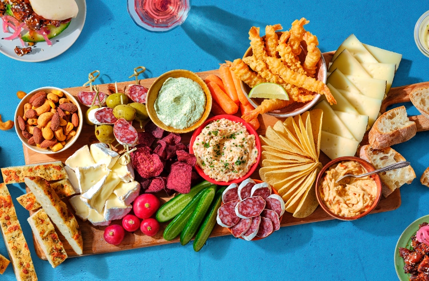 Borrelplank met diverse kazen, worst, groentesticks, toast, noten, smeersels, radijs, komkommer, groentechips, stokbrood en gefrituurde snacks op een blauwe ondergrond