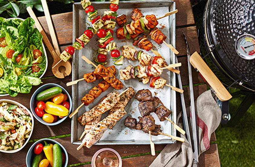 Barbecue met verschillende soorten gegrilde spiesen, verse groene salade, tomaatjes, brood en een pastasalade op een houten buitentafel naast een barbecue.