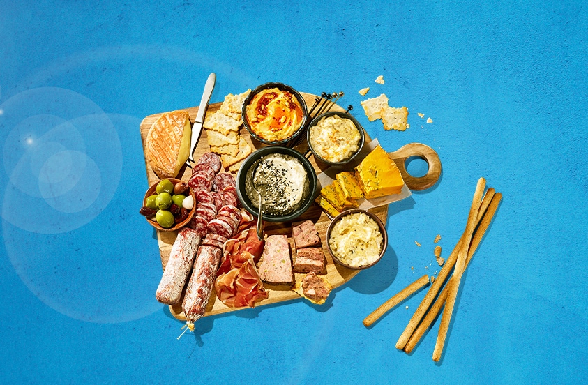 Borrelplank met verschillende soorten vleeswaren, kaas, dips, crackers, olijven en broodstengels op een houten plank, gepresenteerd op een blauwe achtergrond.