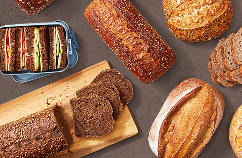 Verschillende soorten volkorenbrood en meergranenbrood, inclusief gesneden brood en sandwiches in een broodtrommel, gepresenteerd op een houten plank en op een tafel.