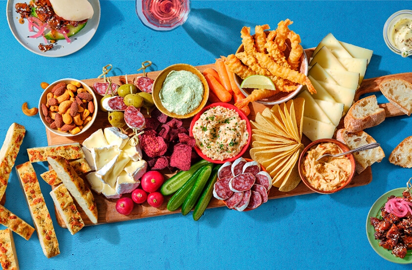 Borrelplank met verschillende kazen, vleeswaren, toastjes, groentesticks, dips, noten, brood, gefrituurde snacks en bijgerechten op een blauwe ondergrond