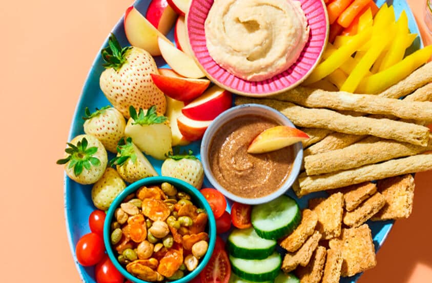 Schaal met verschillende gezonde snacks, waaronder witte aardbeien, appelpartjes, hummus, dip, groenten zoals wortel en gele paprika, broodstengels, komkommer, cherry tomaten, geroosterde peulvruchten en crackers.