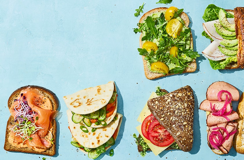Verschillende soorten belegde broodjes, wraps en sandwiches met onder andere zalm, groenten, kaas, avocado en vlees, gepresenteerd op een lichtblauwe ondergrond.