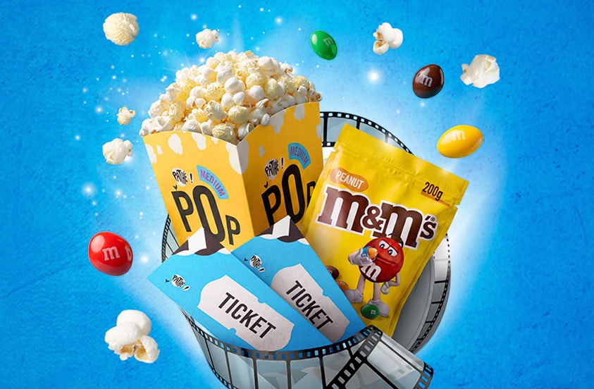 Bak popcorn, zakje peanut M&M's, bioscoopkaartjes en filmrol op blauwe achtergrond, met verspreide stukjes popcorn en M&M's.