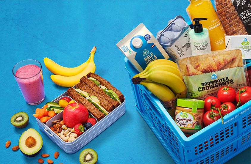 Albert Heijn blauwe winkelmand gevuld met boodschappen zoals brood, croissants, bananen, tomaten, broccoli, kaas, sinaasappelsap, halfvolle melk en handzeep, naast een lunchbox met volkoren sandwiches, fruit, noten en een smoothie op een blauwe ondergrond