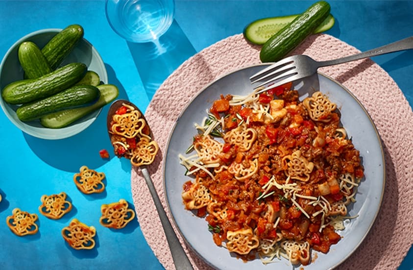 Kindervormpasta met pastasaus en geraspte kaas op een bord, geserveerd met komkommer op een blauw oppervlak