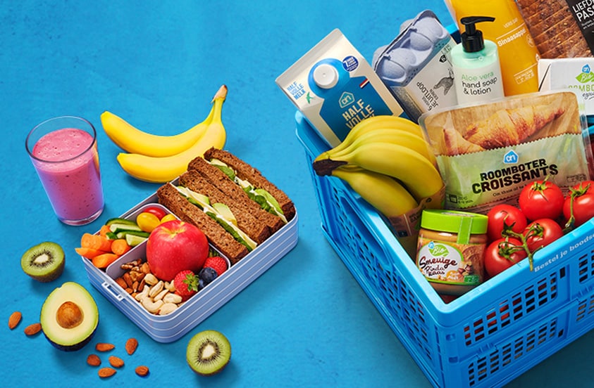 Blauwe boodschappenkrat van Albert Heijn gevuld met diverse verse producten zoals brood, bananen, melk, kaas, broccoli, tomaten, sinaasappelsap, croissants, eieren, handzeep en andere boodschappen; daarnaast een lunchbox met groente, fruit, noten, boterhammen en een glas roze smoothie.