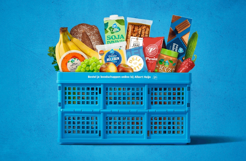 Blauwe Albert Heijn krat gevuld met diverse boodschappen, waaronder vers fruit, brood, kaas, groente, snacks, sojadrink, koffie en verpakte producten tegen een blauwe achtergrond.