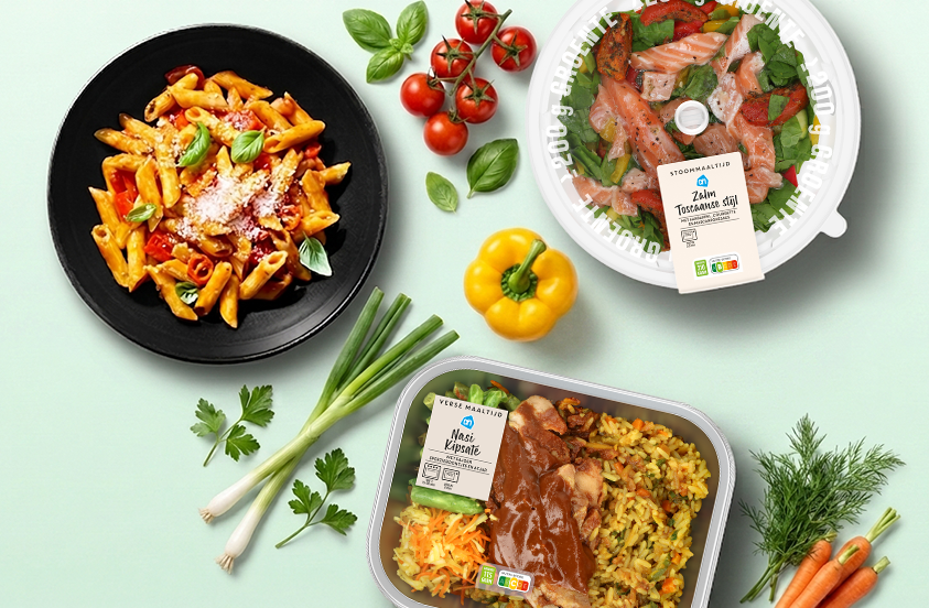 Diverse selectie van verse maaltijden van Albert Heijn, waaronder een bord pasta met tomatensaus en basilicum, een stoombak zalm Toscaanse stijl met groenten, een maaltijdpakket nasi kip sate en verse groenten zoals tomaatjes, paprika, bosui, peterselie en wortels.