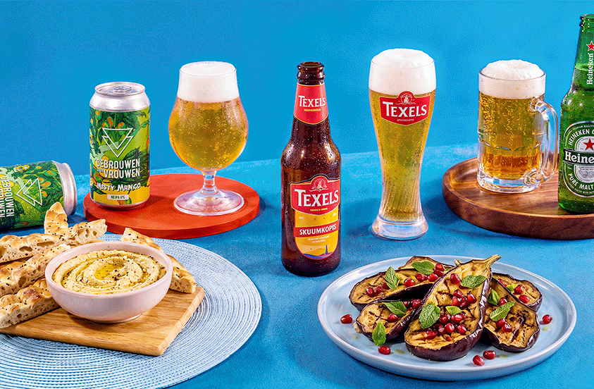 Verschillende bieren van de merken Gebrouwen door Vrouwen, Texels en Heineken gepresenteerd met een glas bier, een bord platbrood met hummus en een bord gegrilde aubergine met granaatappelpitten op een blauwe achtergrond.