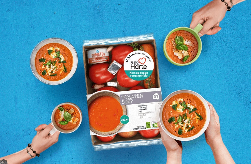 Voedselpakket van Albert Heijn voor tomatensoep met verse tomaten, tomatenpuree en bouillonblokjes, omringd door vier kommen tomatensoep die door verschillende handen worden vastgehouden tegen een blauwe achtergrond