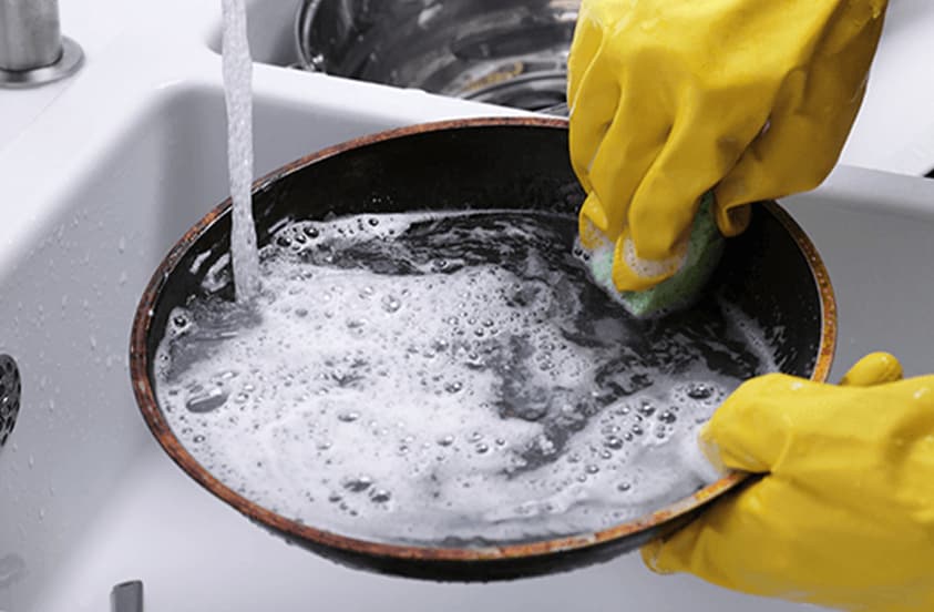 Iemand met gele schoonmaakhandschoenen wast een koekenpan met een spons onder stromend water in een gootsteen.