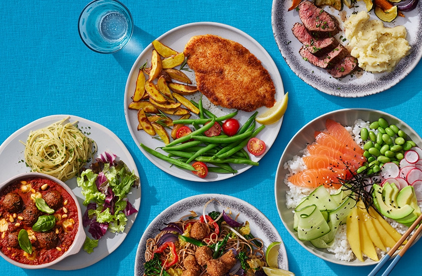 Verschillende kleurrijke borden met gerechten, waaronder een salade met zalm en avocado, pasta met gehaktballetjes, schnitzel met gebakken aardappelen en groenten, en een bord met biefstuk, aardappelpuree en groenten, geserveerd op een blauwe achtergrond.