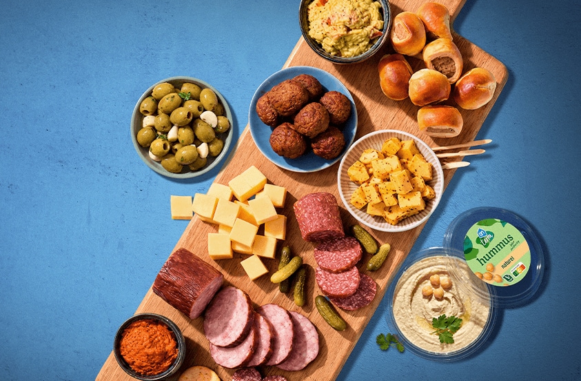 Borrelplank met verschillende hapjes, waaronder kaasblokjes, plakjes worst, mini augurken, olijven, gehaktballetjes, broodjes met vulling, hummus van het merk AH en diverse dips, gepresenteerd op een houten plank tegen een blauwe achtergrond.