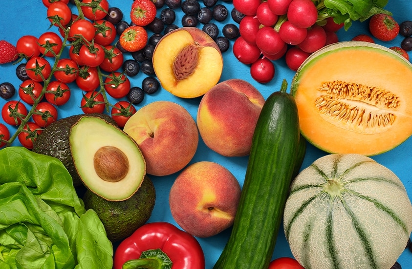 Verse groenten en fruit, waaronder rode paprika, avocado, tomaten, citroen, sla, perziken en bessen op een blauwe achtergrond.
