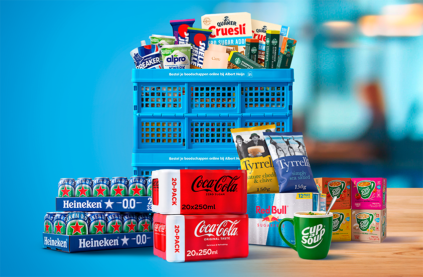 Afbeelding van diverse A-merk producten zoals Heineken 0.0 bier, Coca-Cola, Red Bull, Quaker Cruesli, Tyrrells chips, Cup-a-Soup, Alpro en andere snacks en dranken, gepresenteerd met een blauwe krat van Albert Heijn op een houten tafel.